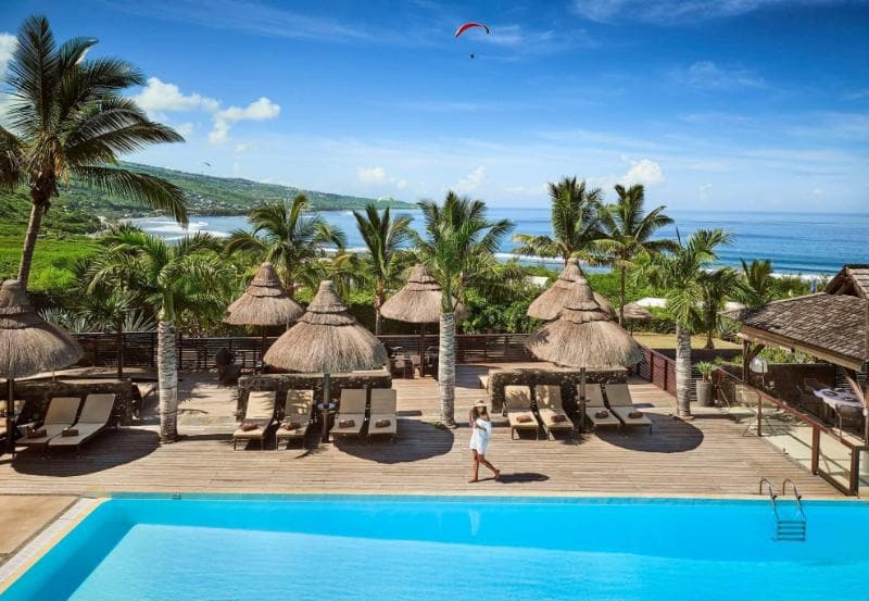 The pool at Iloha Seaview in Saint-Leu with thatched parasols, palms, the ocean and a paraglider in the sky
