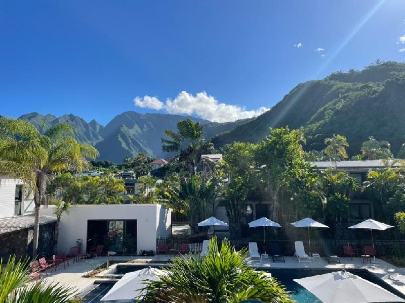 The Dimitile Hôtel & Spa pool with white parasols among palm trees, green mountains and blue sky behind