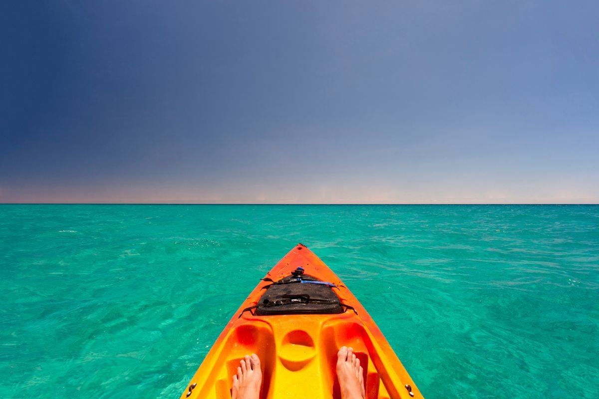 Vue à la première personne depuis l'avant d'un kayak orange flottant sur une eau turquoise cristalline, pieds visibles, ciel bleu profond