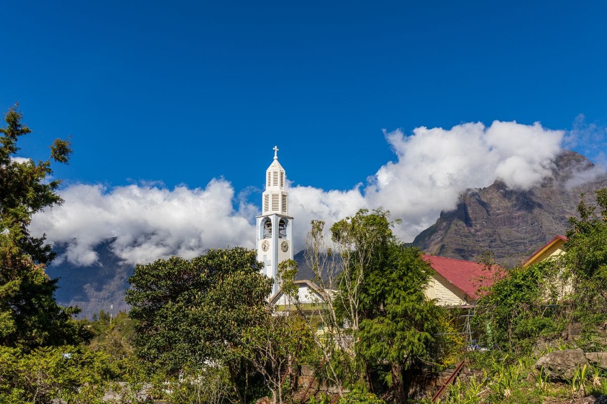 The white bell tower of Cilaos church under a blue sky, with the cirque walls in the clouds