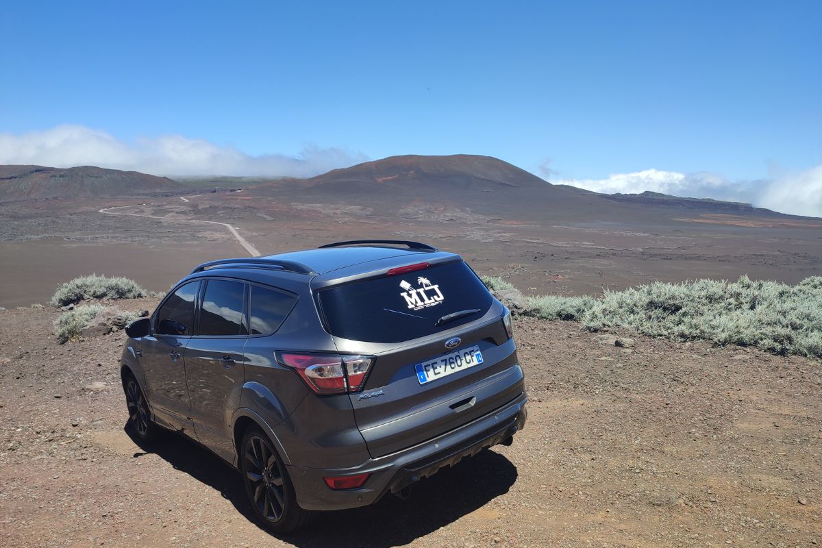 SUV Ford Kuga gris MLJ Concept vu de l'arrière dans le paysage minéral de la Plaine des Sables au Piton de la Fournaise, sticker MLJ Concept visible sur la lunette, ciel bleu profond