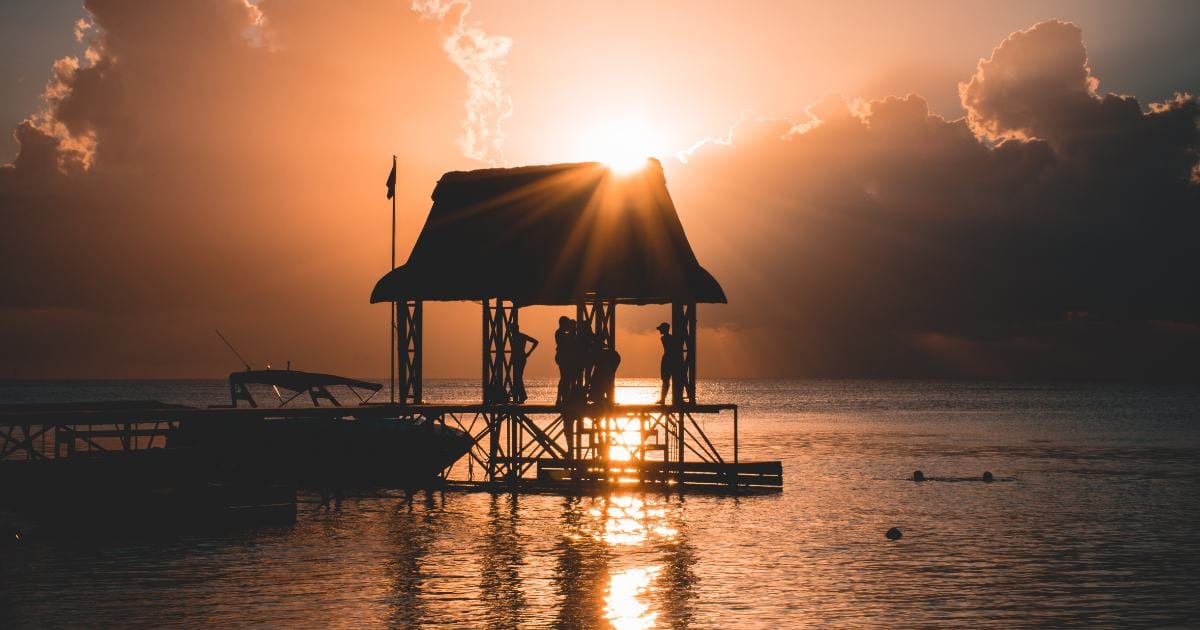 A thatched kiosk at the end of a jetty, figures silhouetted against the sunset