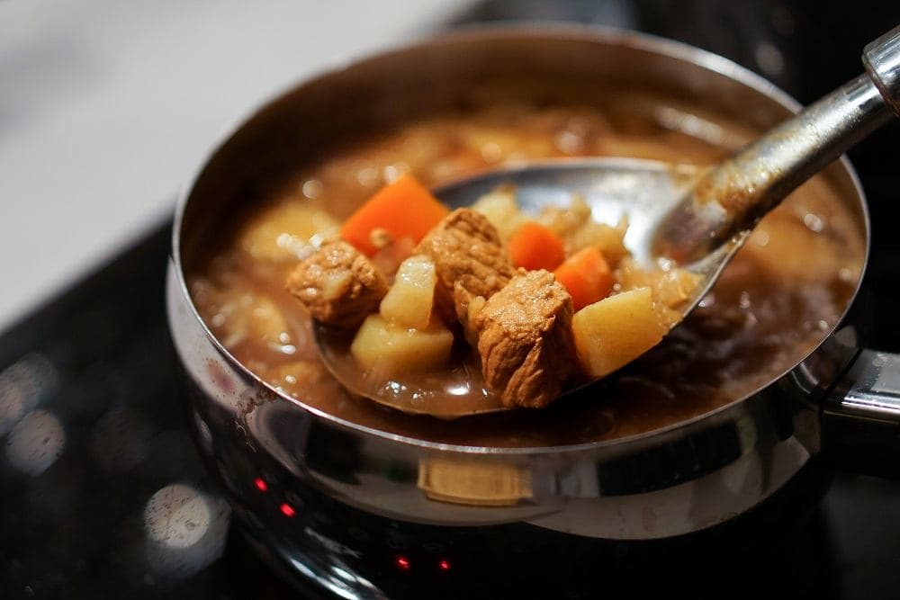Louche soulevant un carry créole en cuisson, morceaux de viande, carottes et pommes de terre dans une sauce orangée