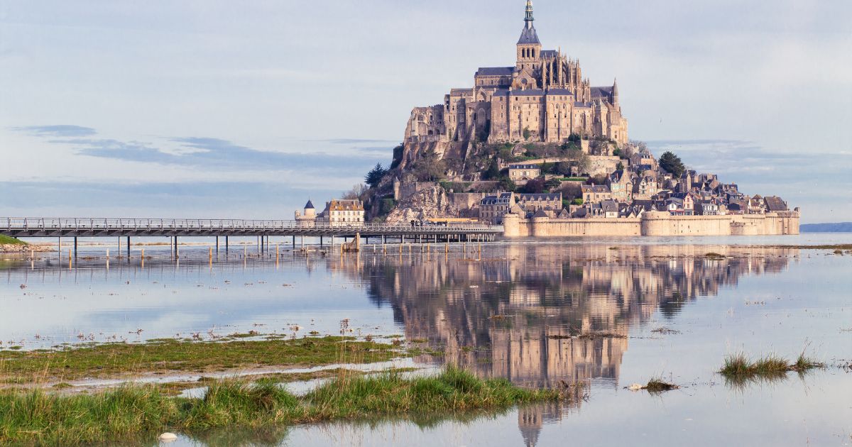 Le Mont-Saint-Michel se reflète dans la baie à marée haute, ciel pastel et passerelle au premier plan