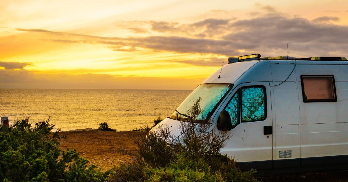 Van blanc aménagé stationné face à la mer au coucher du soleil sur une côte rocheuse, ciel orangé, ambiance road trip libre