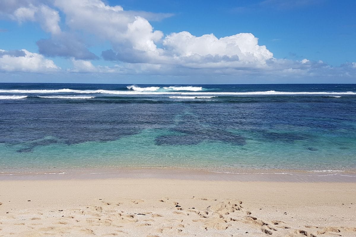 Lagon peu profond aux eaux claires, vagues brisées sur la barrière de corail au loin, sable au premier plan