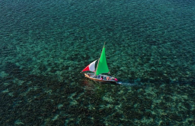 A traditional outrigger with a green and red sail on a turquoise Mauritian lagoon, coral visible below