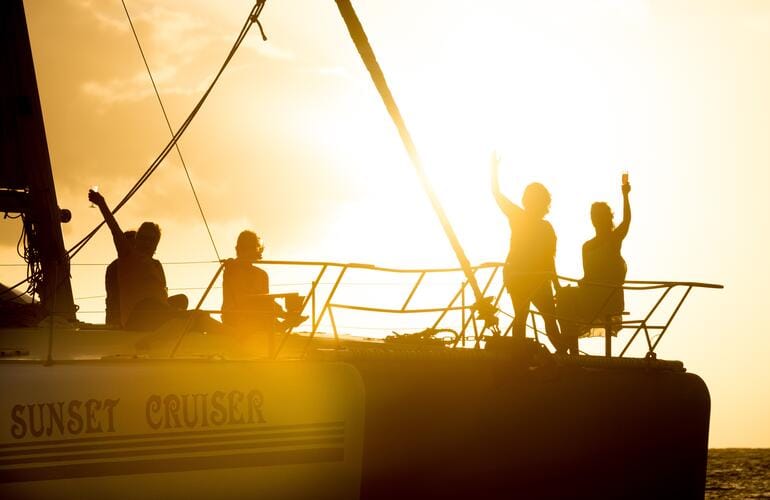 Silhouettes de passagers bras levés sur un catamaran au coucher du soleil