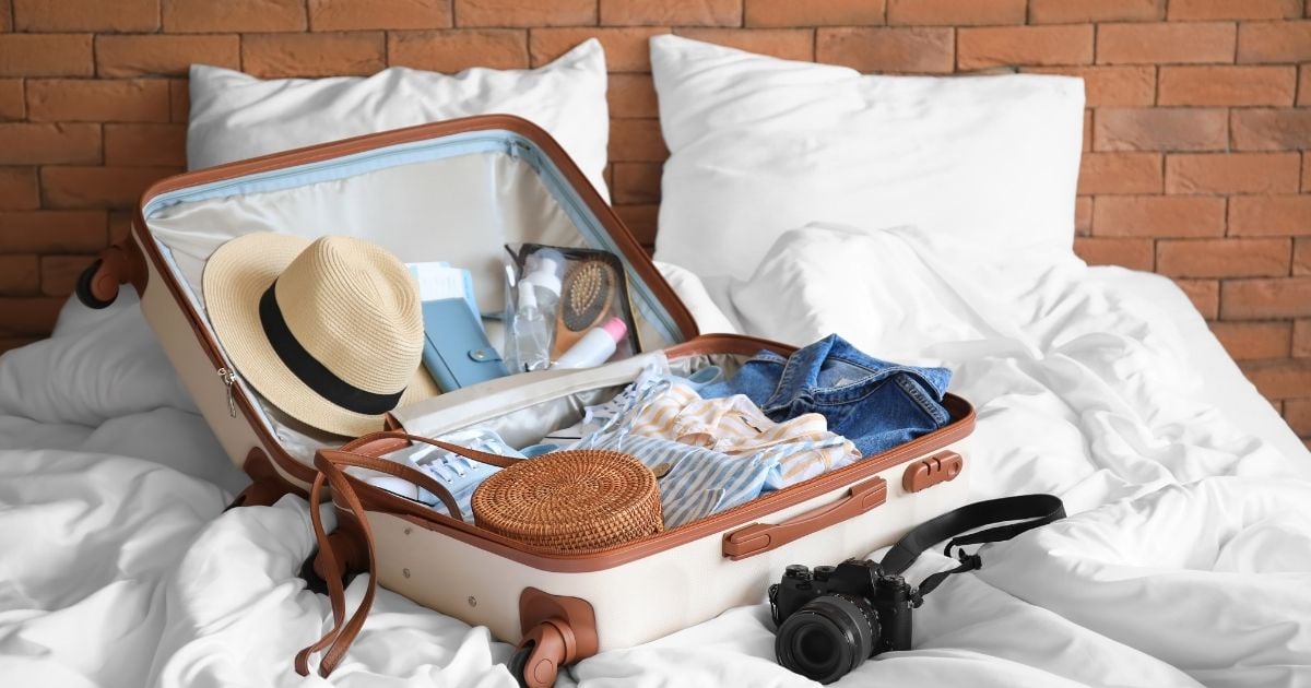 A hard-shell suitcase open on a bed, containing a straw hat, clothes, a camera and a toiletry bag: typical preparations for a tropical trip