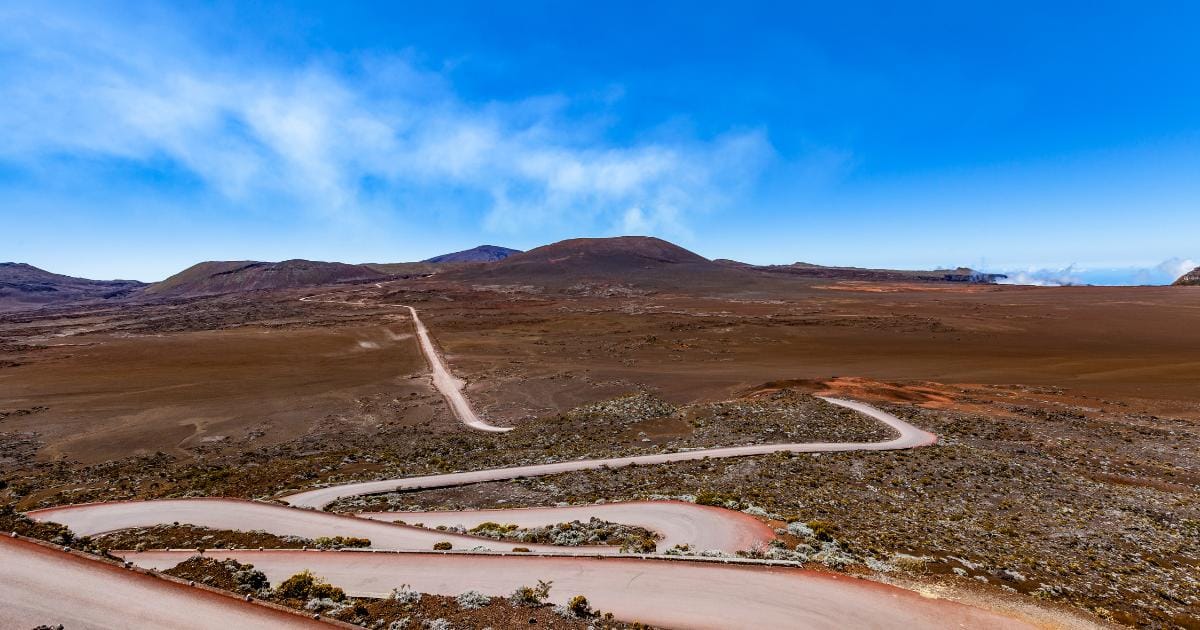 Route en lacets de la Plaine des Sables avec sols ocre rouge et lointaines fumerolles volcaniques