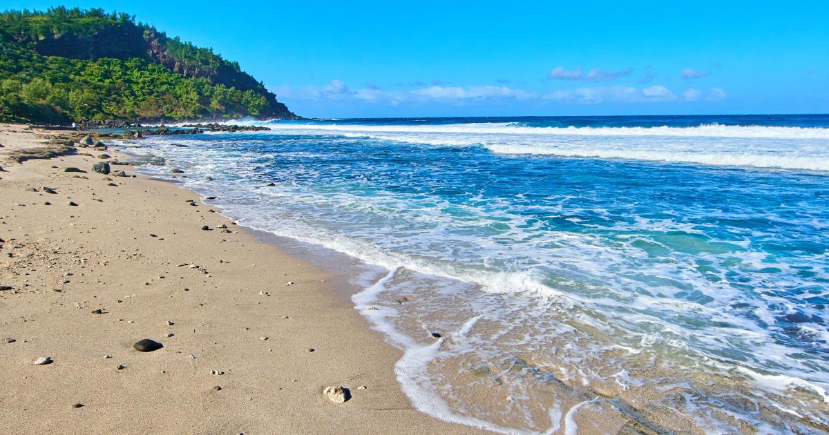 Plage de sable clair de la côte ouest de La Réunion en mars : mer turquoise, vagues sur la barrière de corail et falaise verdoyante en arrière-plan