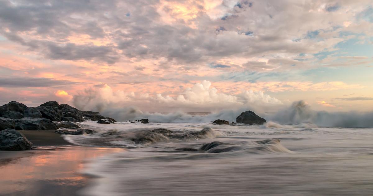 Wild coast of Reunion Island in February at sunset: black sand beach, basalt rocks and waves under a pink sky heavy with austral summer