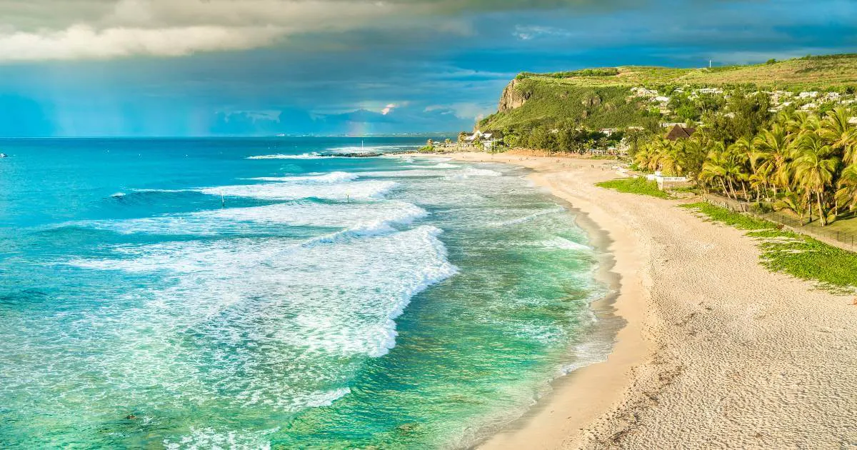 A west coast beach in Reunion Island in April: light sand, turquoise lagoon, coral reef and a green cliff under an austral autumn sky