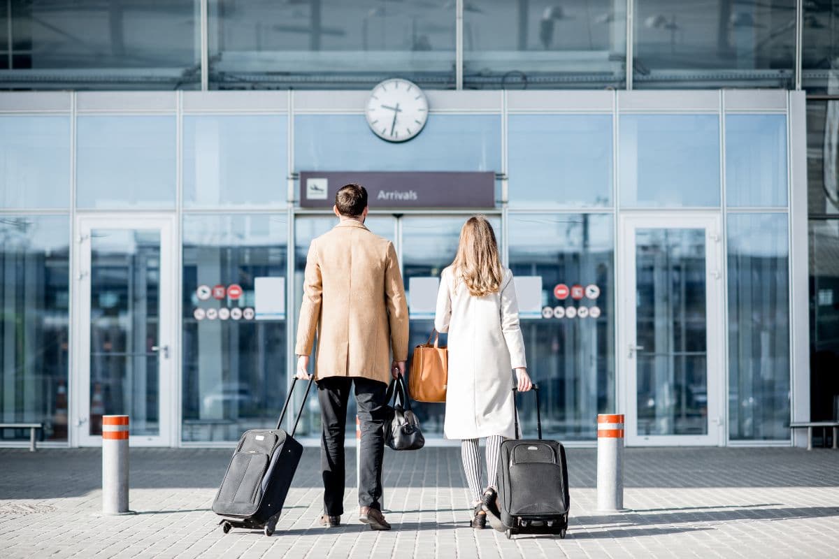 Couple tirant des valises à roulettes devant l'entrée vitrée d'un terminal d'aéroport, sous le panneau des arrivées