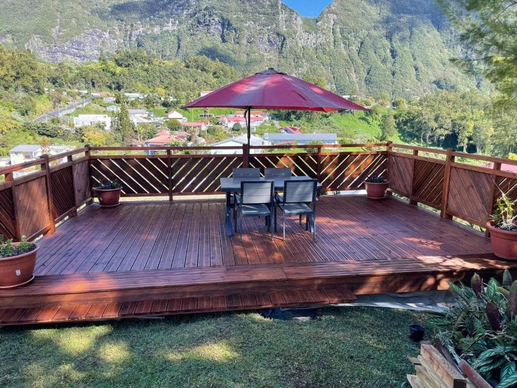 Terrasse en bois avec table et parasol rouge, face au village de Grand-Îlet et aux remparts de Salazie
