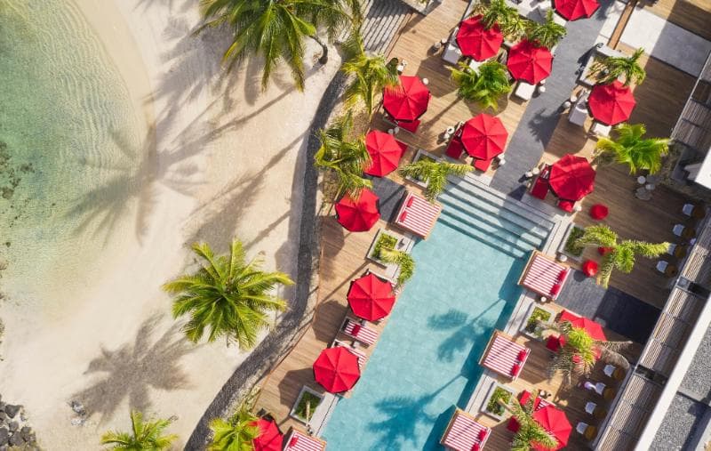 Vue aérienne du LUX* Grand Baie : longue piscine bordée de parasols rouges et de palmiers, au ras de la plage