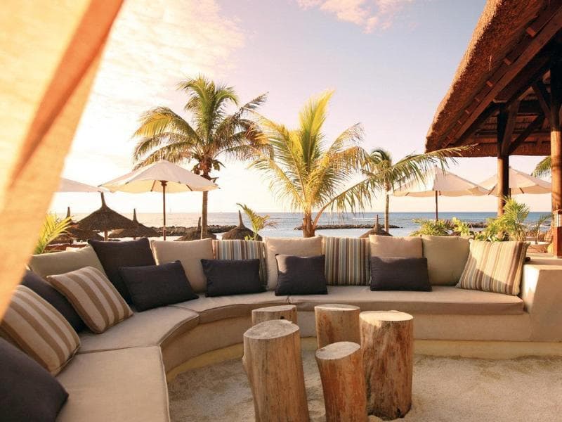 Outdoor lounge at Veranda Pointe aux Biches: a sofa with striped cushions, tree-trunk stools under a thatched roof and the beach at sunset