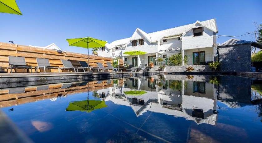 Façade blanche de l'hôtel Les Géraniums se reflétant dans la piscine, parasols verts et transats
