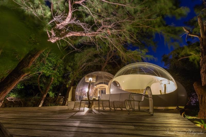 Deux bulles transparentes éclairées de nuit sur une terrasse en bois, sous les arbres de l'île aux Cerfs